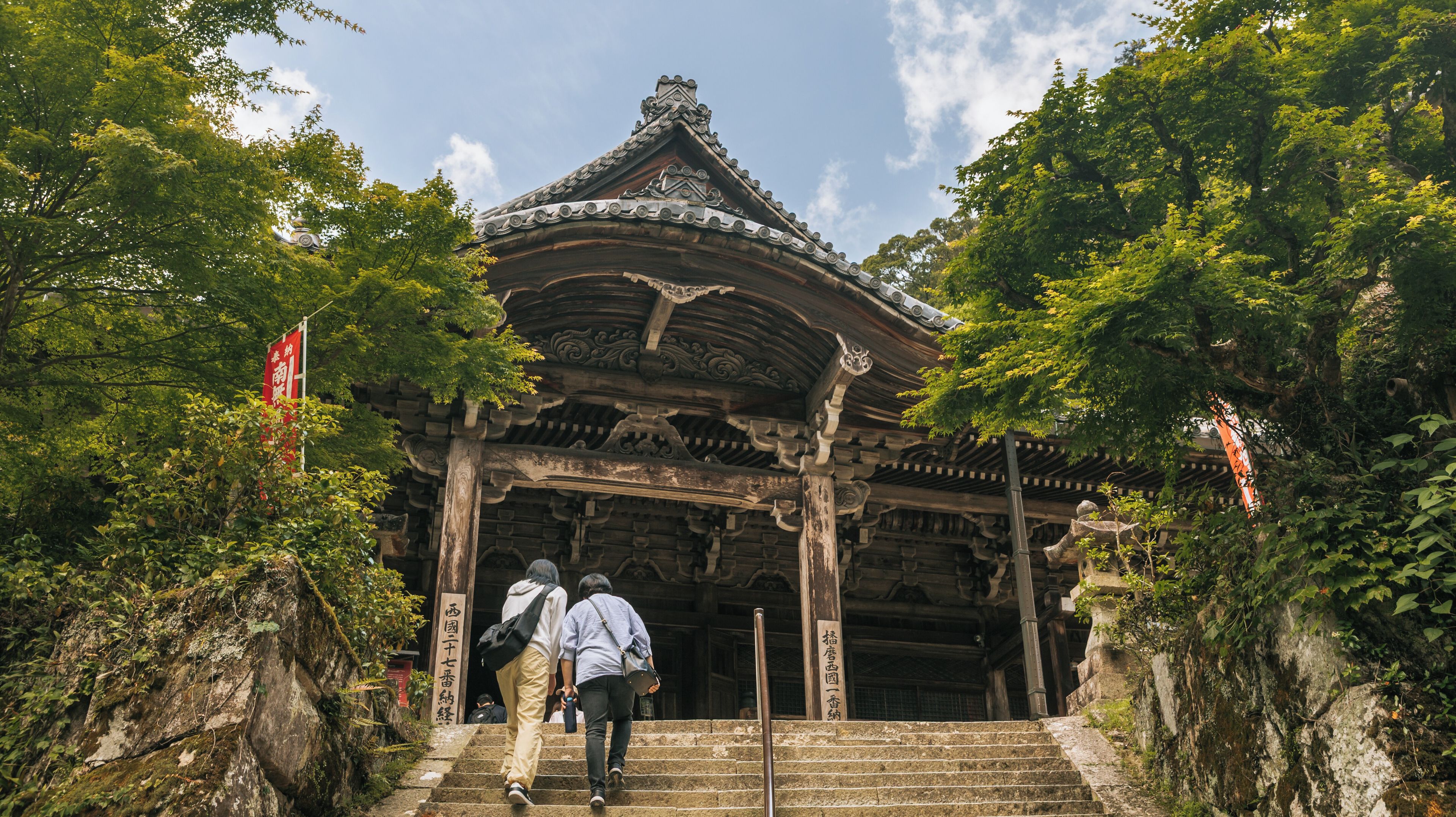 Entrada tradicional d'un temple japonès amb arquitectura de fusta ornamentada, escales de pedra i vegetació exuberanta.