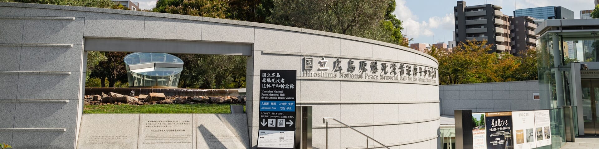 A memorial building with a curved entrance, signage in multiple languages, and a glass structure in the background.