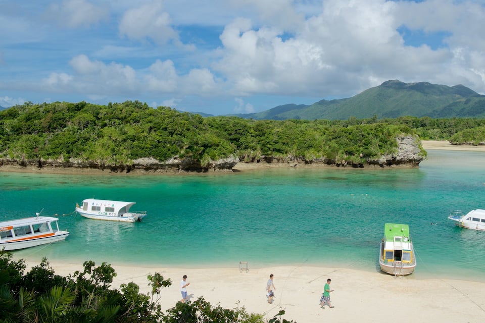 A beach with boats, clear turquoise water, and lush green hills in the background.