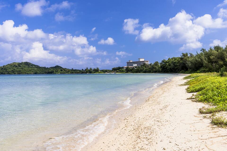 A beach with clear turquoise water, a sandy shore, and a building on a hill in the background.