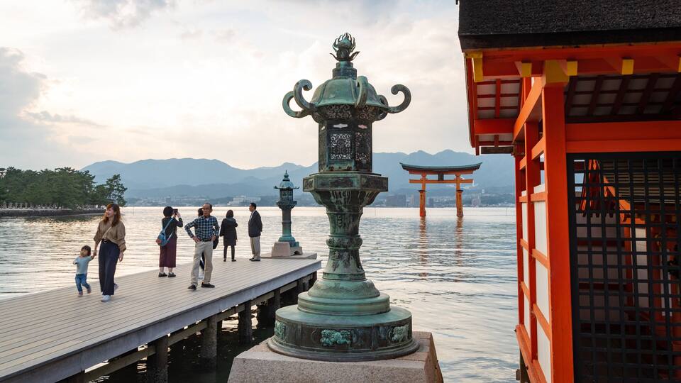 A wooden pier with people walking, a large bronze statue, and a red torii gate in the background.