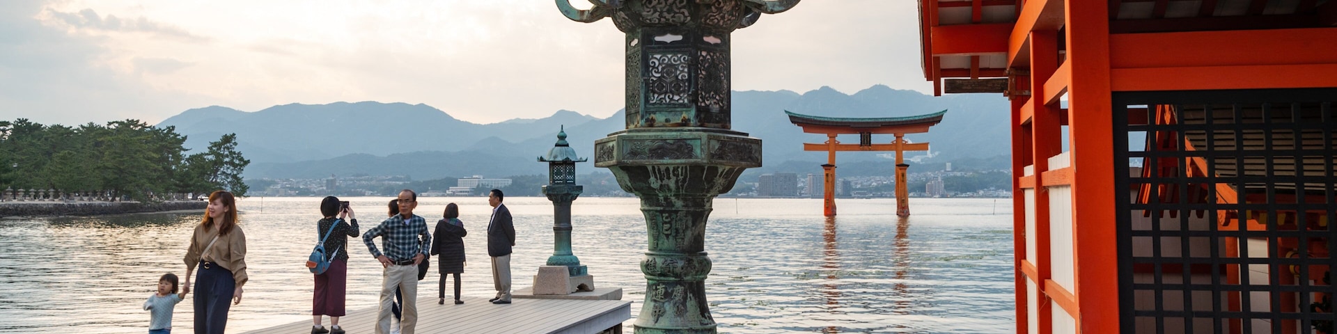 Un muelle de madera con gente caminando, una gran estatua de bronce y un torii rojo al fondo.