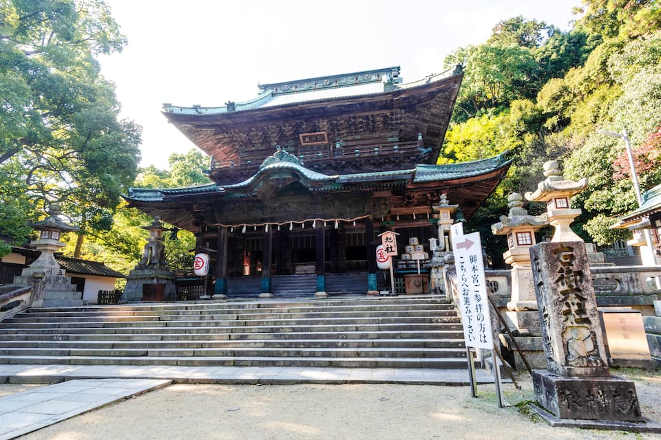 Un templo japonés tradicional con un techo grande y ornamentado, escalones de piedra que conducen a la entrada y un letrero con caracteres japoneses.