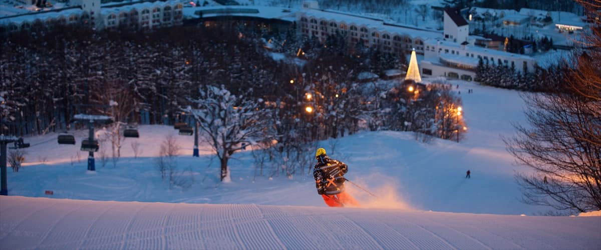 A snowy ski slope with a skier in motion, surrounded by snow-covered trees and buildings in the background.