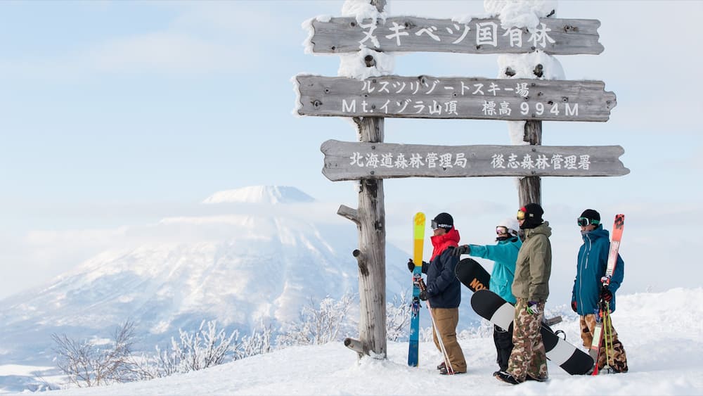 Un groupe de skieurs au sommet d’une montagne, près d’un panneau indiquant le lieu et l’altitude.