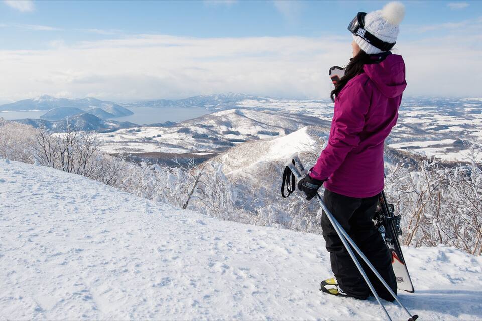 A person skiing on a snowy mountain with a panoramic view of snow-covered hills and a lake in the distance.
