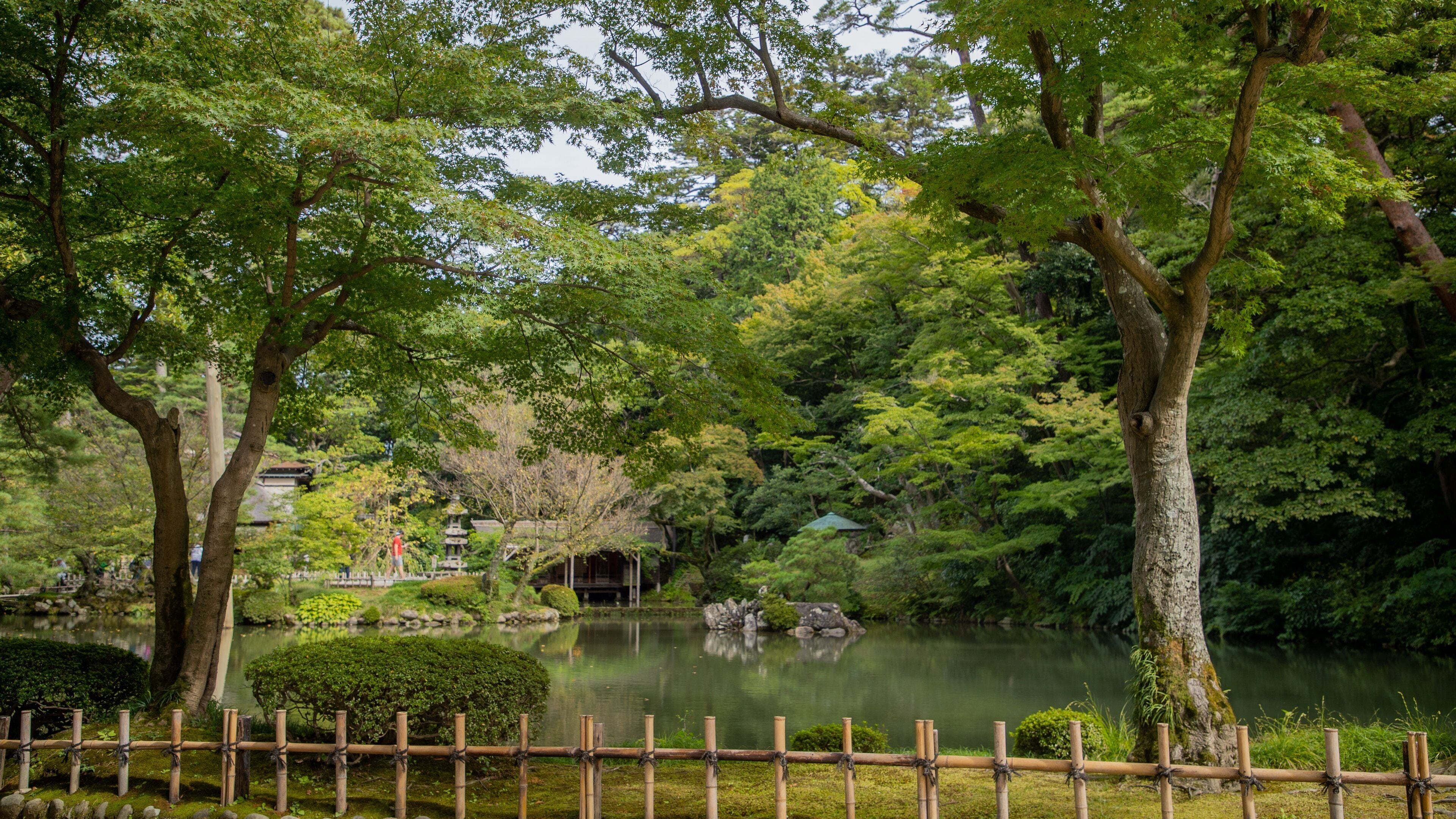 A serene Japanese garden with a pond, bamboo fence, and traditional buildings.