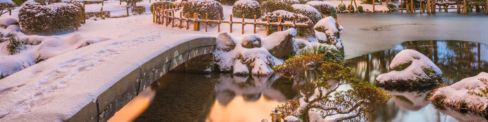 Un jardín cubierto de nieve con un puente, árboles y un estanque.