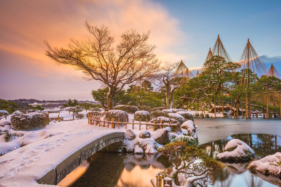 A snow-covered garden with a bridge, trees, and a pond.