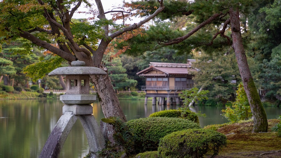 Un farol de piedra, una edificación japonesa tradicional y un estanque en un jardín tranquilo.