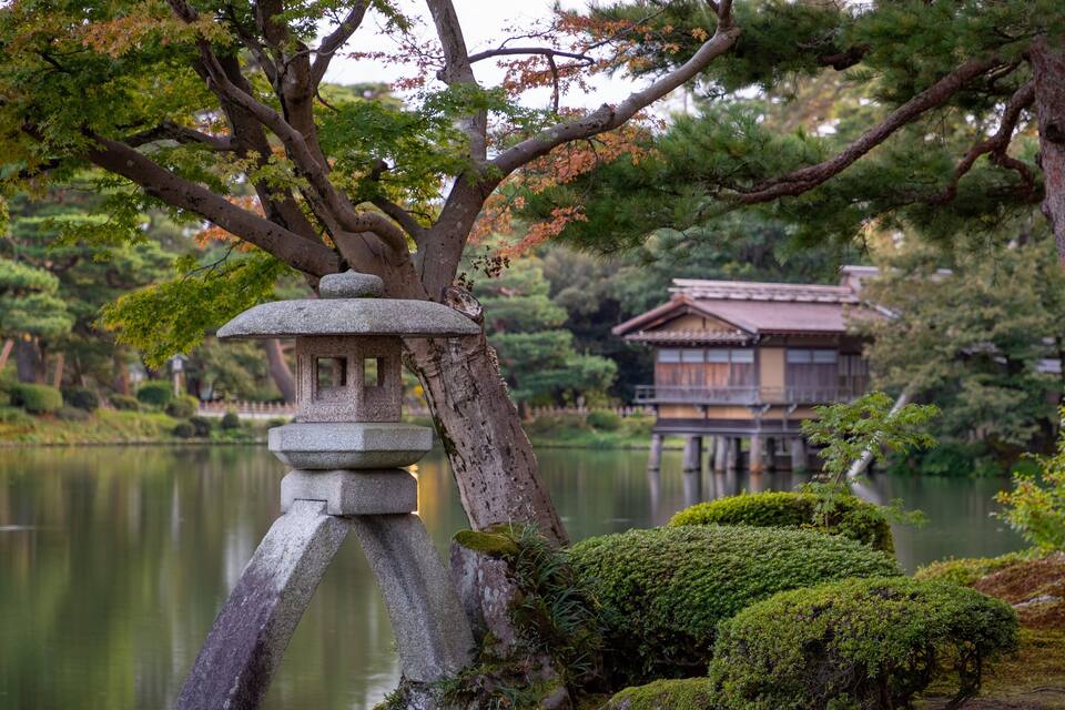 Um lampião de pedra, um edifício japonês tradicional e um lago num jardim tranquilo.