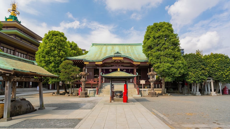 A traditional Japanese temple with a green roof, stone lanterns, and a large tree in the foreground.