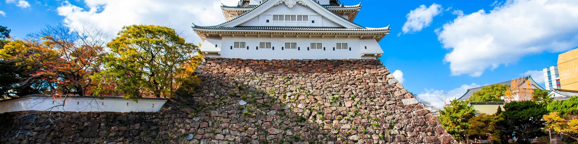 A traditional Japanese castle with multiple tiers, a stone base, and a clear blue sky with clouds.