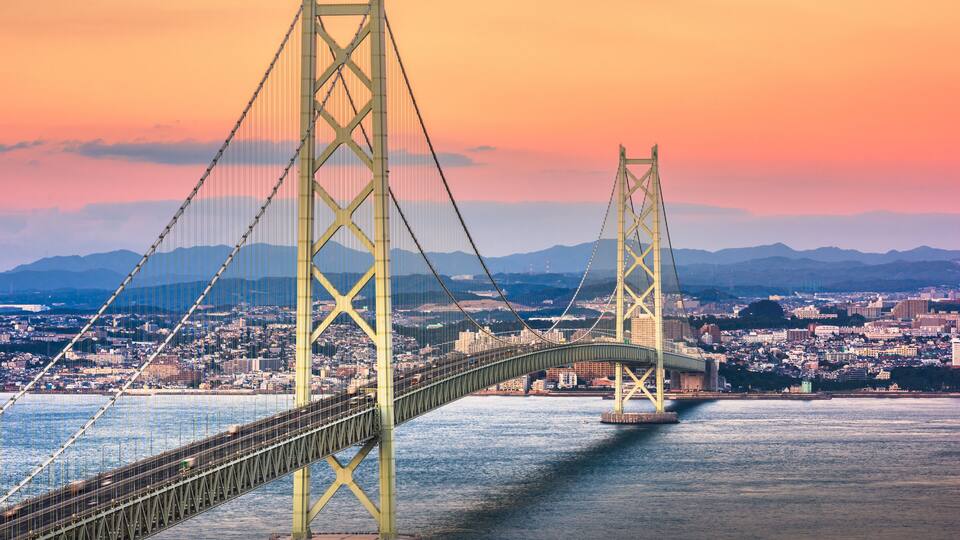 Uma ponte suspensa com duas torres, atravessando um corpo de água, com um horizonte urbano ao fundo.