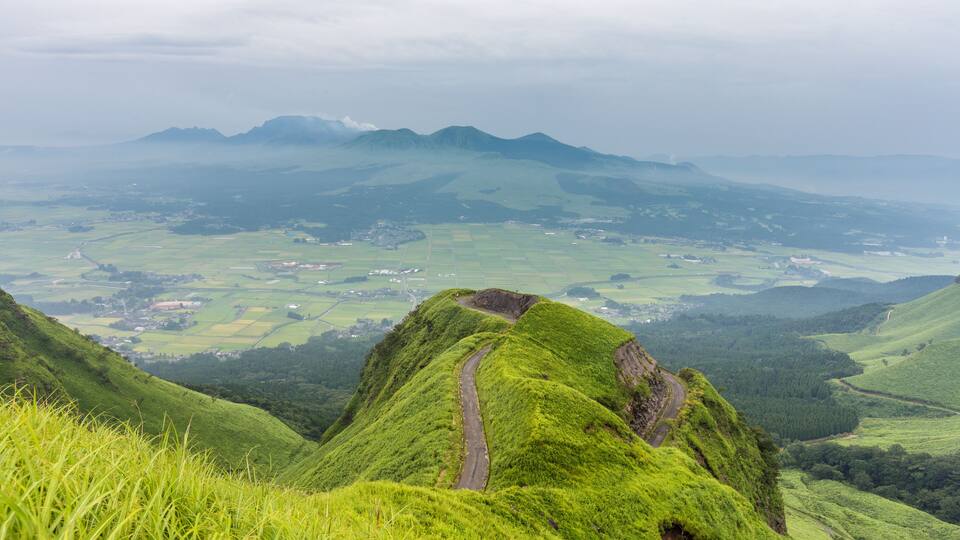 A lush green valley with a winding road, rolling hills, and distant mountains.