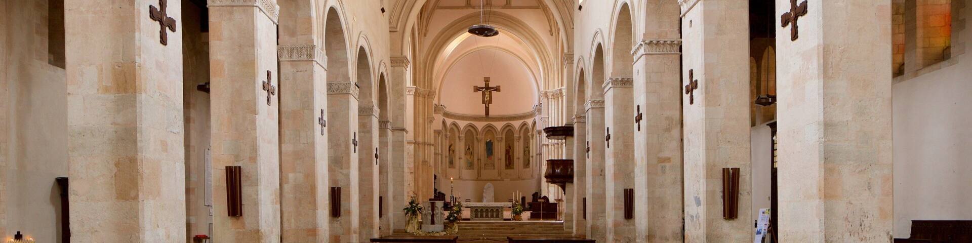 Interior de iglesia con hileras de bancas de madera, techos abovedados y múltiples columnas de piedra con cruces.