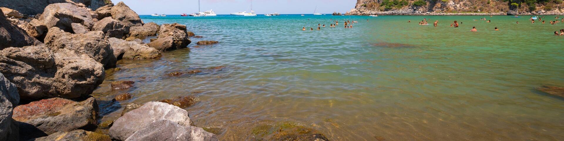 A rocky coastline with clear turquoise water, boats, and people swimming.