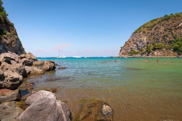 A rocky coastline with clear turquoise water, boats, and people swimming.
