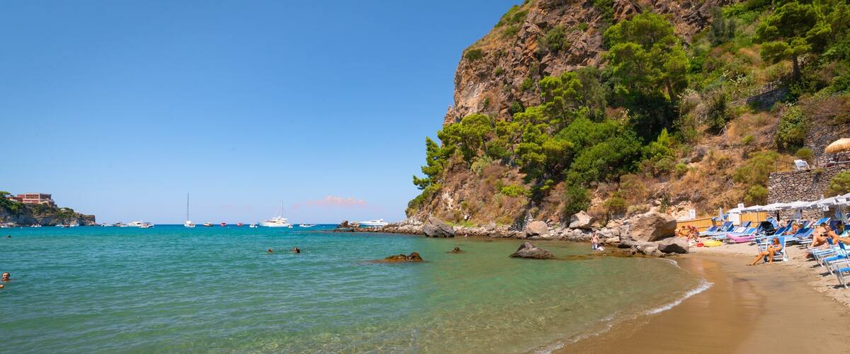 A beach with clear turquoise water, a rocky cliff with greenery, and a few boats in the distance.