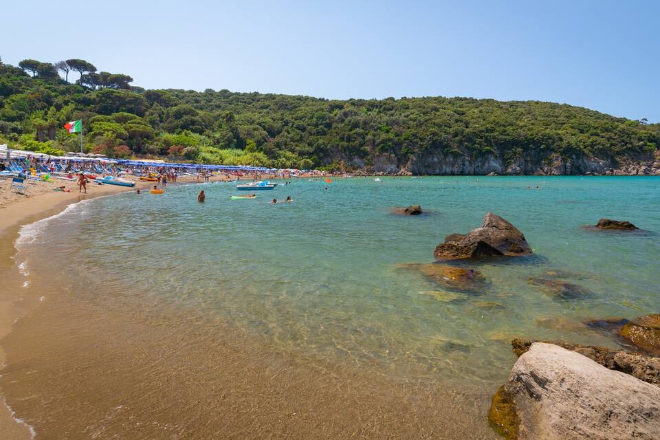 A beach with clear turquoise water, rocky outcrops, and a crowd of people under umbrellas.