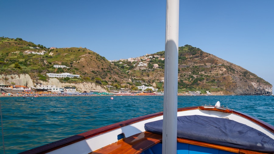 Un barco sobre aguas marinas tranquilas, con un pueblo costero y colinas al fondo.