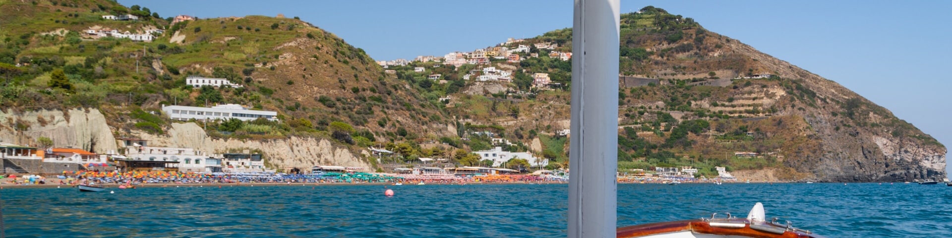 Um barco em águas calmas do mar, com uma vila costeira e colinas ao fundo.