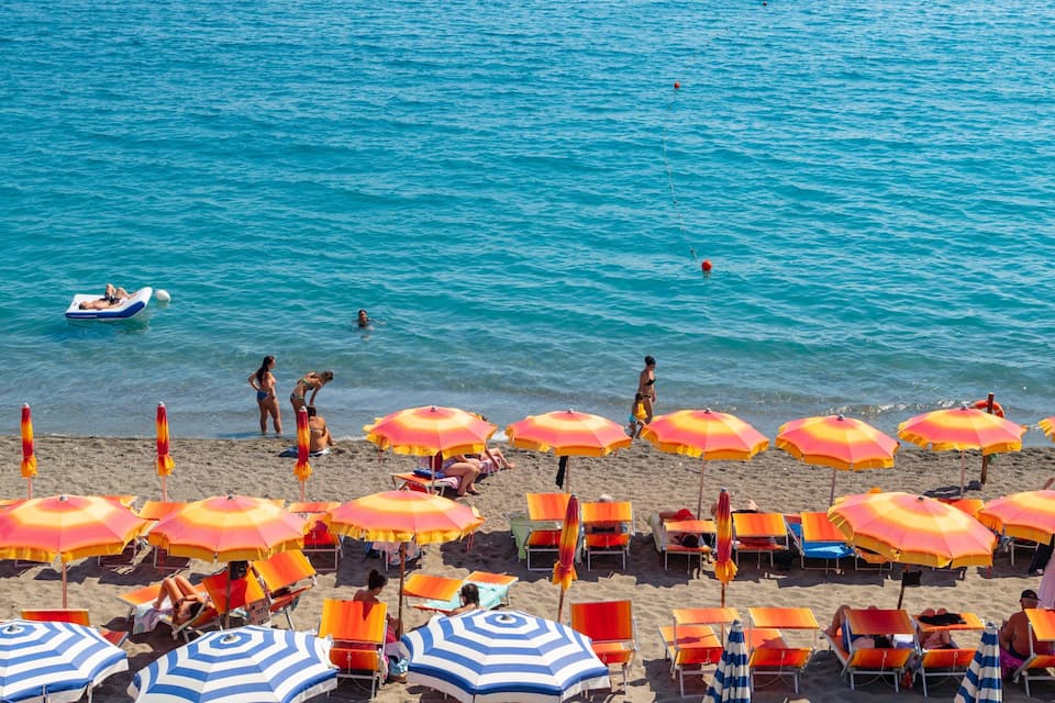 A beach with clear blue water, people swimming, and numerous colorful umbrellas and sun loungers.