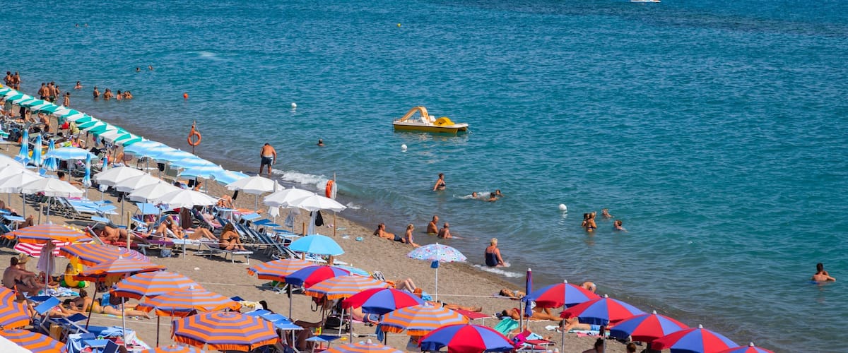 A crowded beach with people swimming and sunbathing under colorful umbrellas.