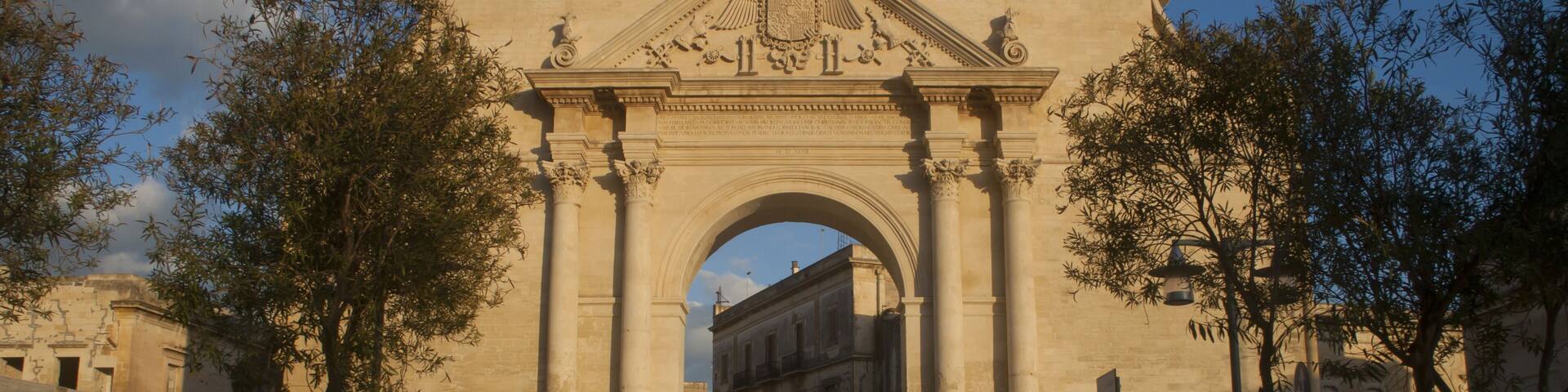 An ornate stone archway with classical columns and a crest, surrounded by trees and benches.