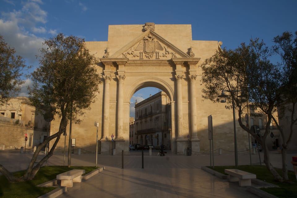 An ornate stone archway with classical columns and a crest, surrounded by trees and benches.