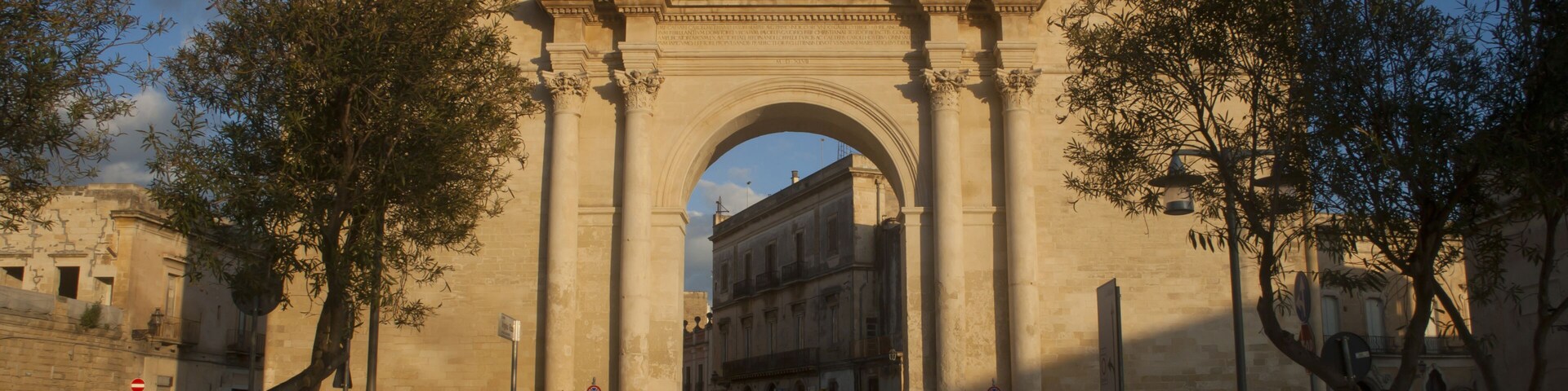Un arco de piedra ornamentado con columnas clásicas y un escudo, rodeado de árboles y bancos.