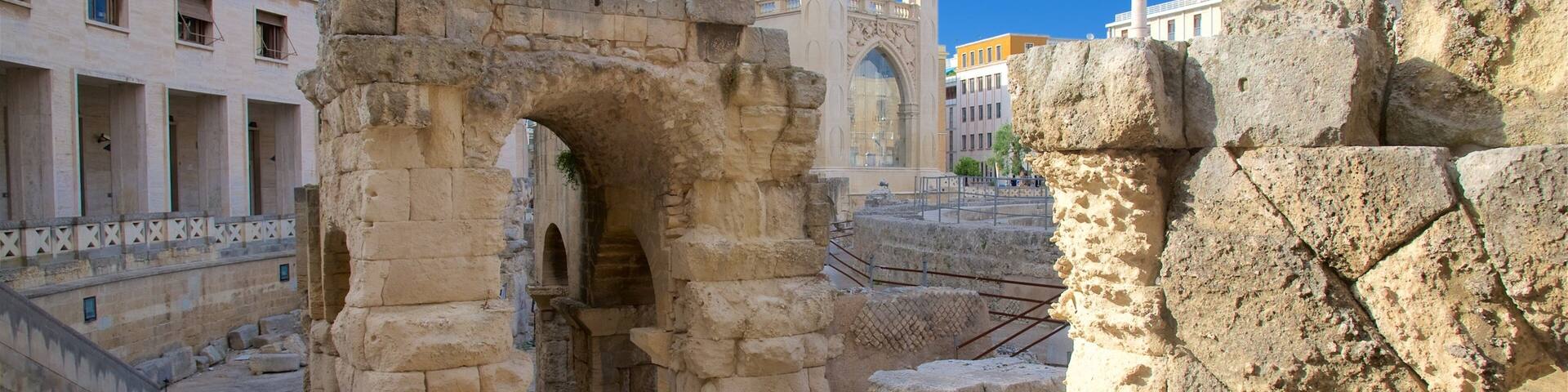 Ancient ruins with stone arches and columns, a modern building in the background, and a statue on a pillar.