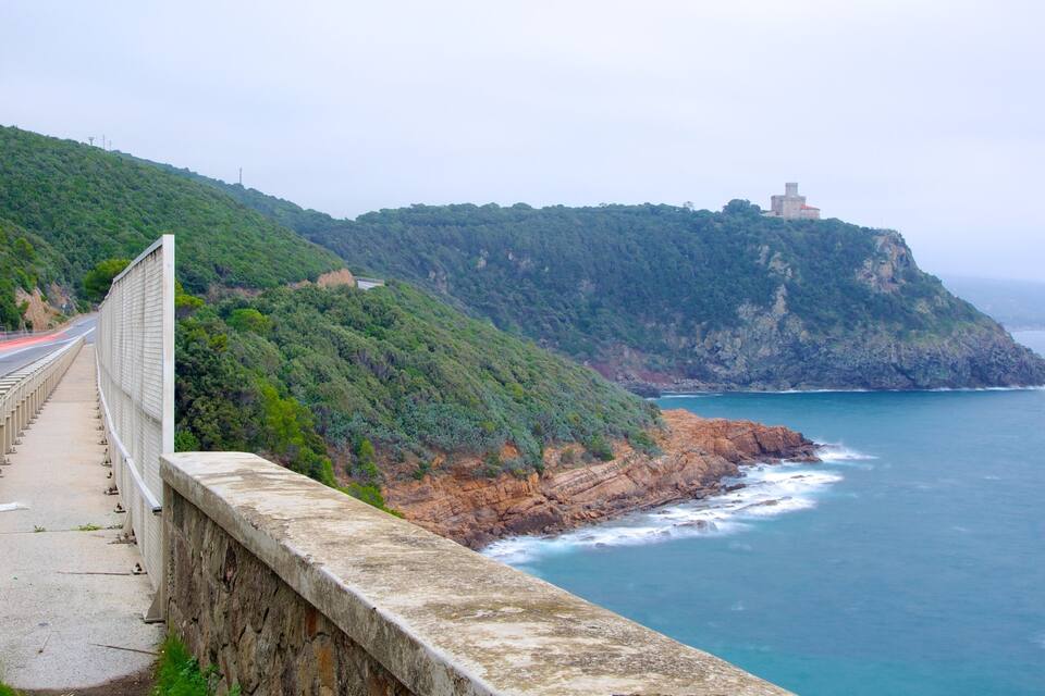 A coastal road with a guardrail, a rocky cliff, and a lighthouse on a hill.