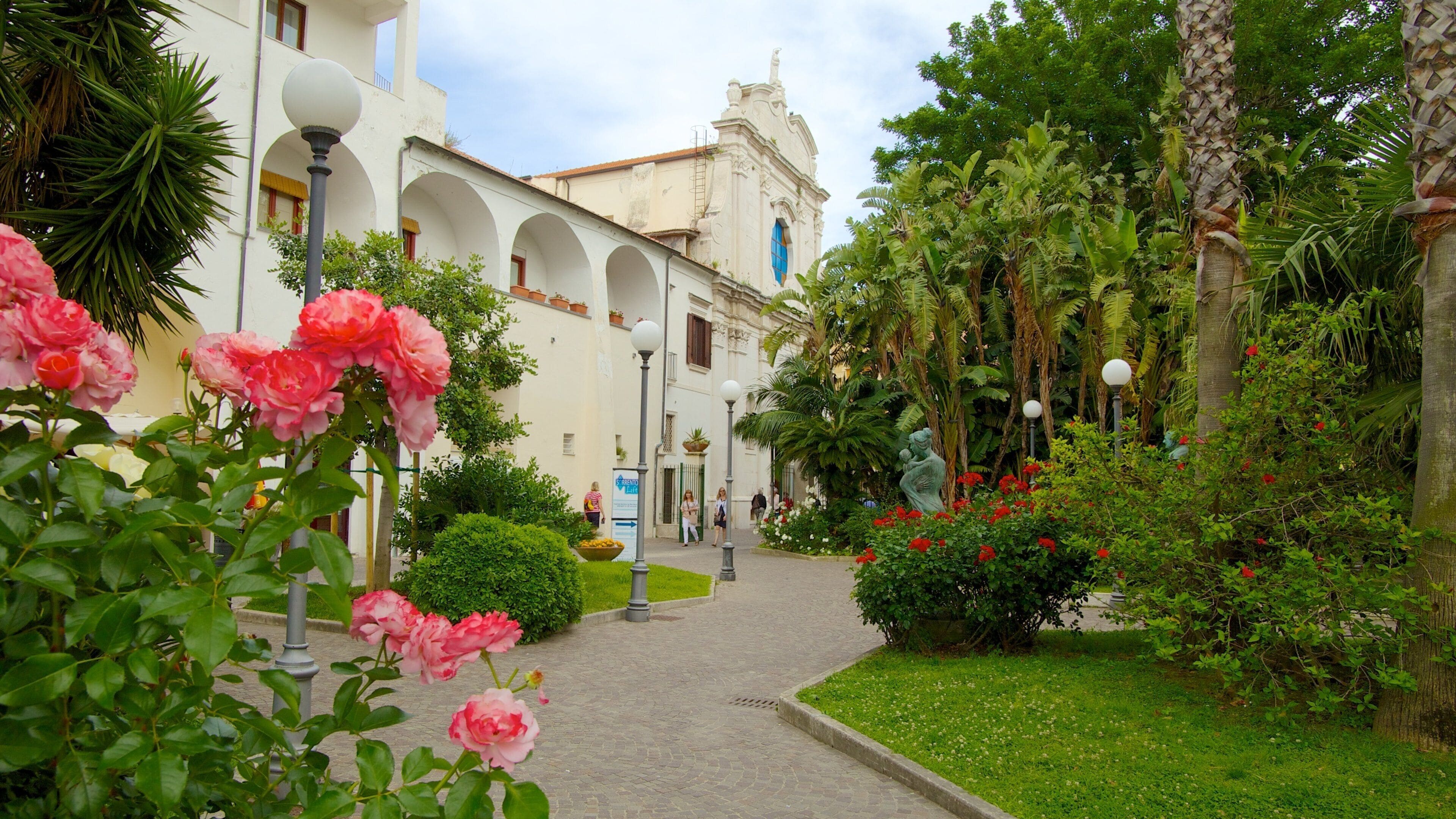 A garden with pink roses, a white building with arches, and a church tower.