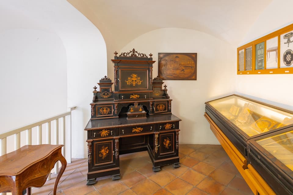 A room with a dark wooden cabinet with ornate carvings, a glass display case with artifacts, and a framed painting on the wall.