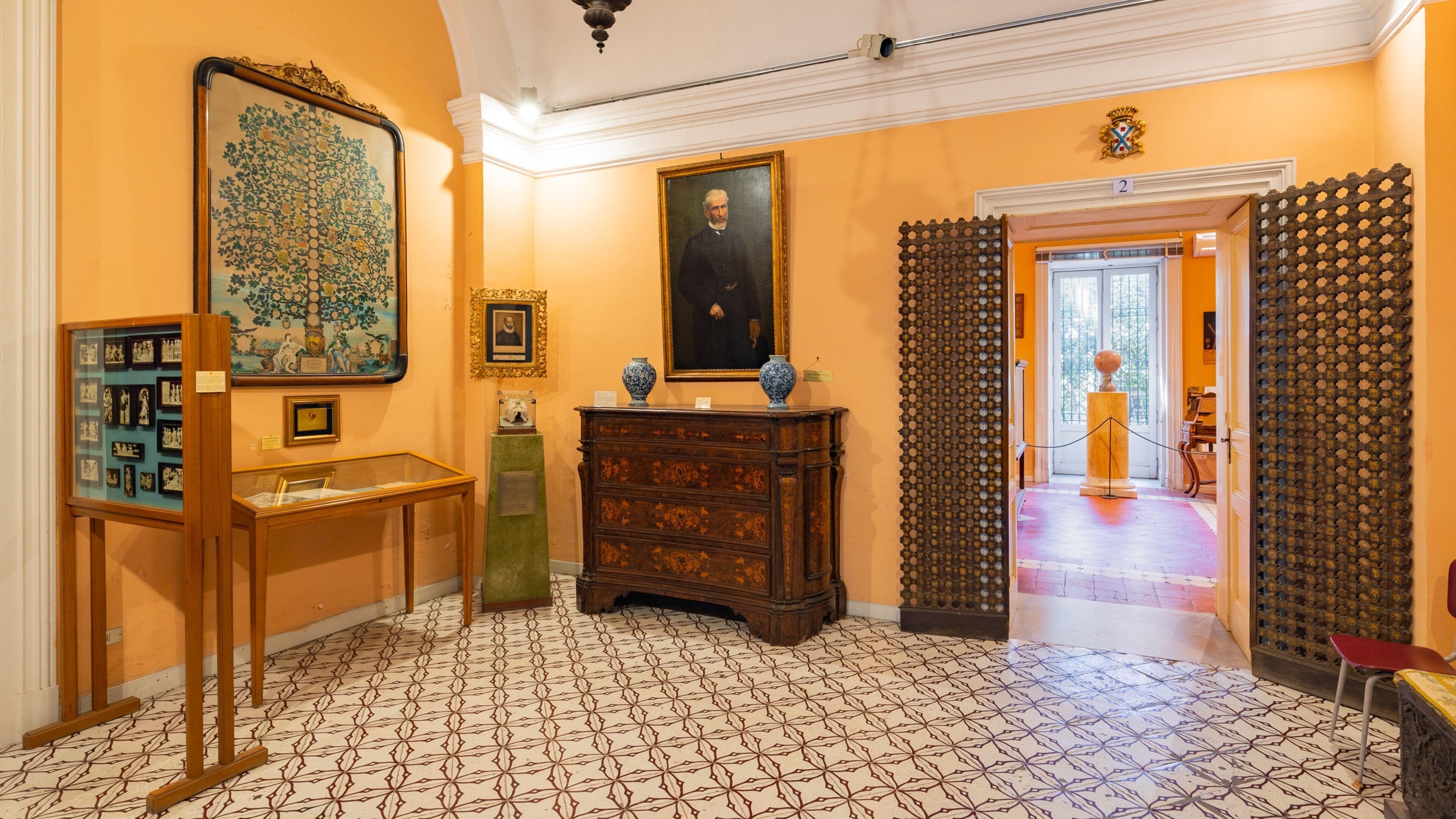 A hallway with patterned floor tiles, a large framed painting, a wooden cabinet, and a door with a decorative grille.