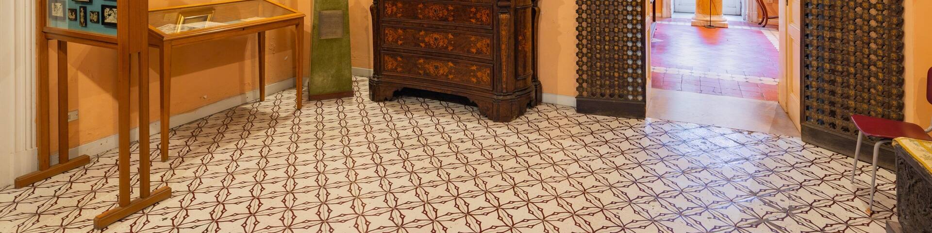 A hallway with patterned floor tiles, a large framed painting, a wooden cabinet, and a door with a decorative grille.