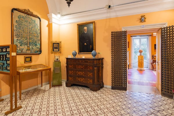 A hallway with patterned floor tiles, a large framed painting, a wooden cabinet, and a door with a decorative grille.