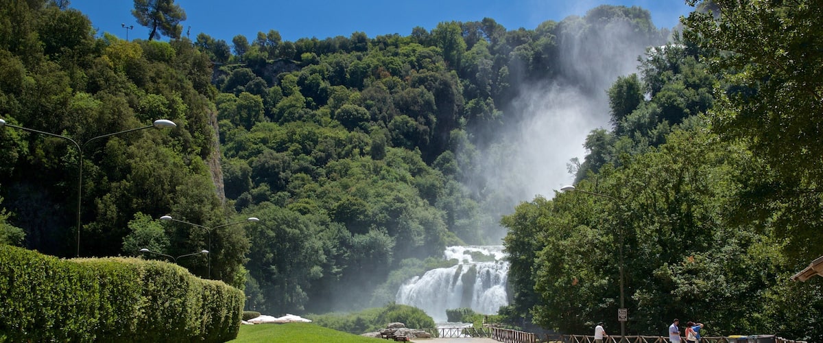 Uma cascata rodeada por vegetação exuberante e um caminho pavimentado com bancos.