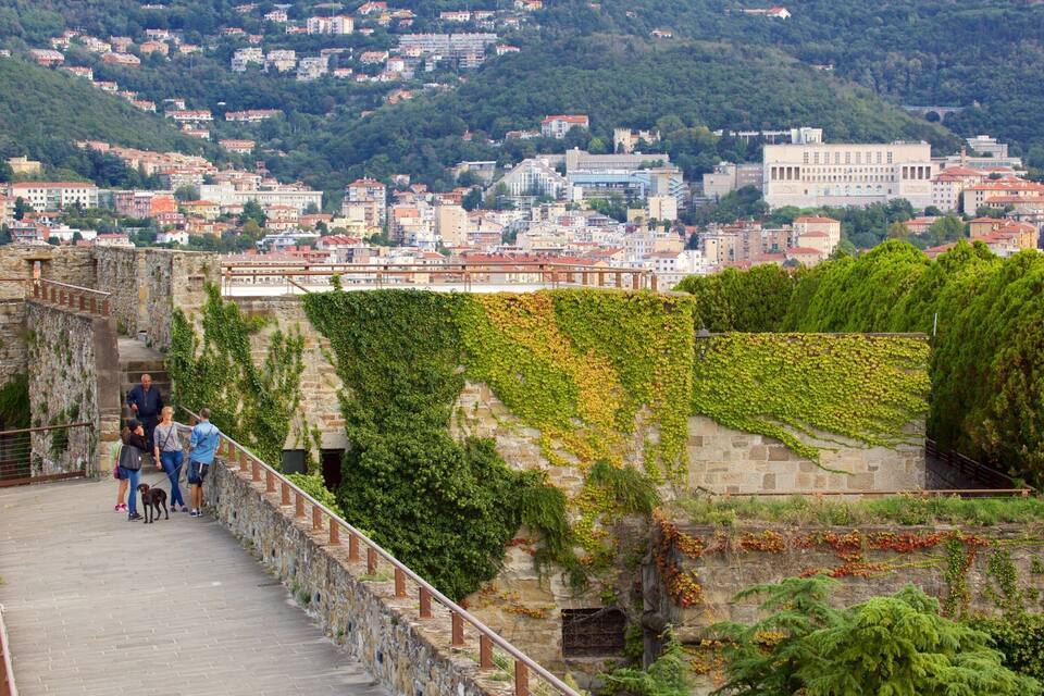 A stone bridge with greenery, a cityscape in the background, and people walking.