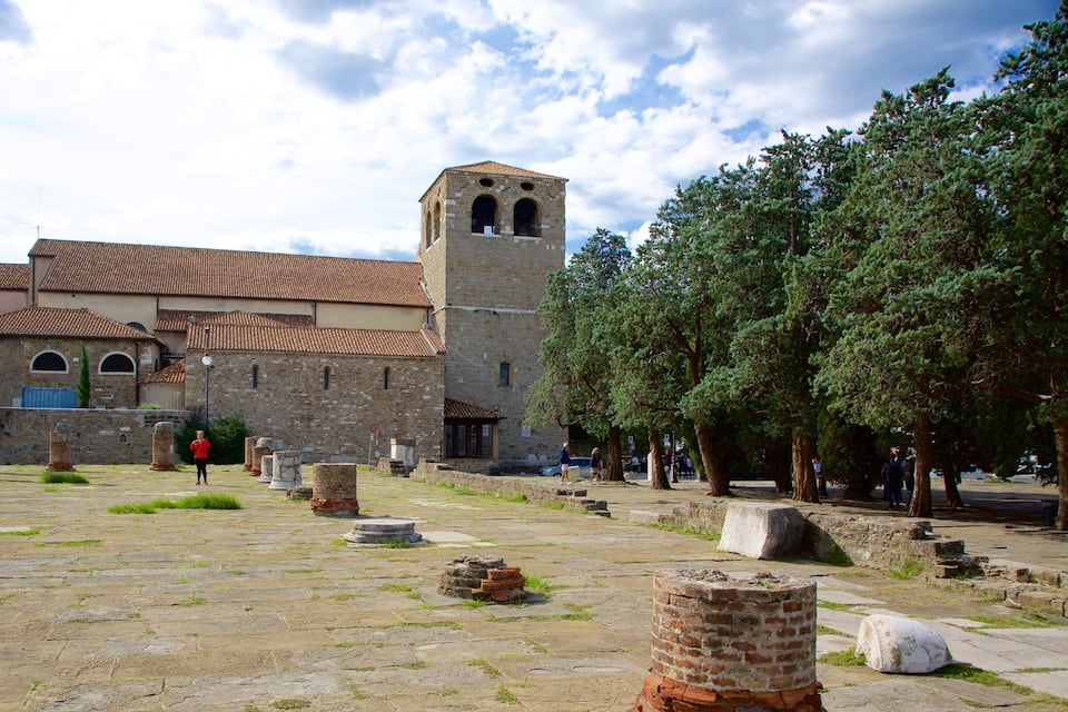 Um pátio antigo de pedra com ruínas, uma igreja com um campanário e árvores.