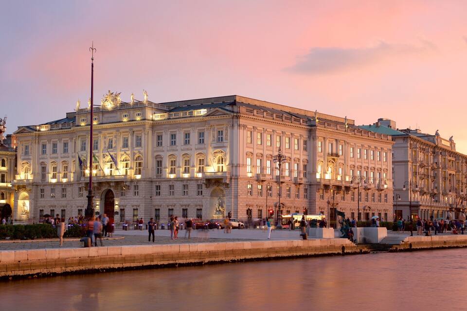 A large, ornate building with classical architecture, a waterfront, and people walking along the promenade.