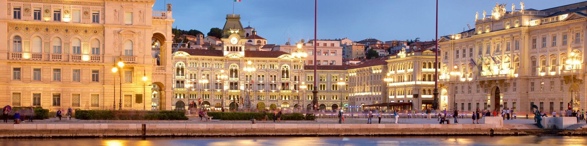 A riverside promenade with historic buildings, a prominent clock tower, and a statue in the center.