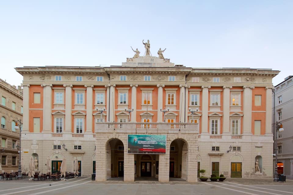 A historic building with classical architecture, featuring a central entrance with arches, a large statue above the entrance, and a banner with text.