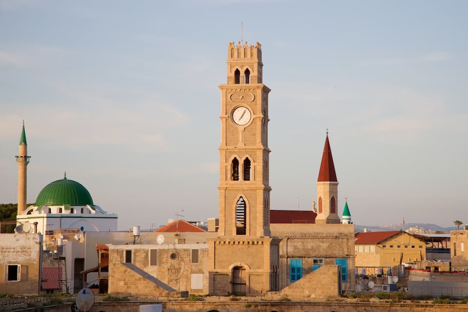 A historic cityscape with a prominent clock tower, a mosque with a green dome, and a church with a red spire.