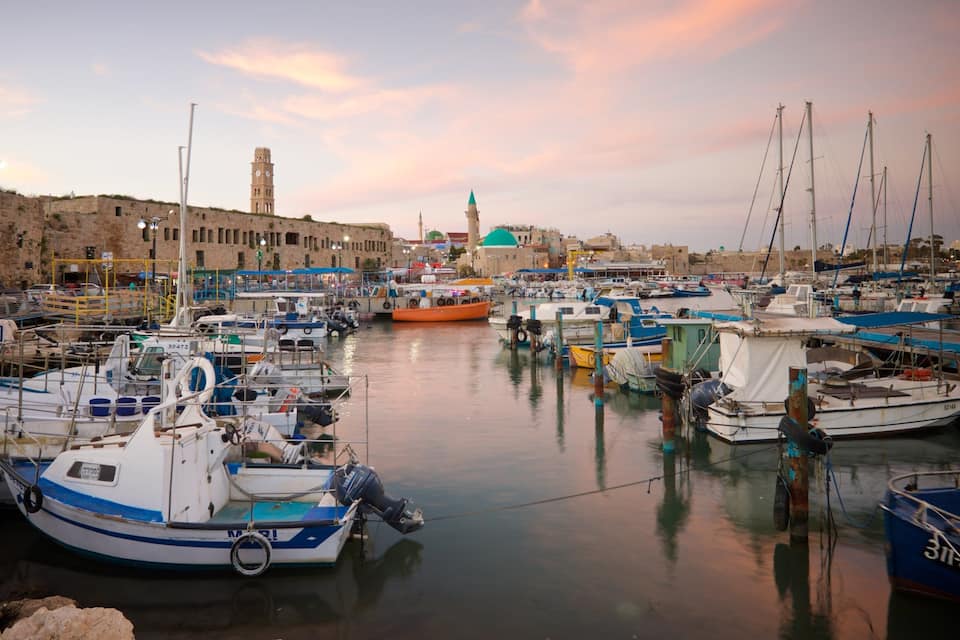 A harbor with numerous boats docked, a prominent clock tower, and a mosque with a green dome in the background.