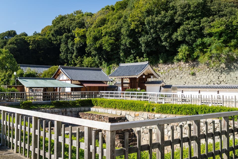 Traditional Japanese buildings with tiled roofs, surrounded by lush greenery and a stone wall.