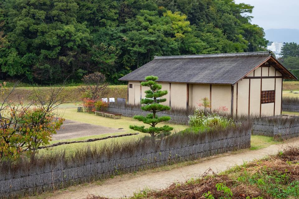 Un bâtiment traditionnel japonais, entouré d’une zone boisée et bordé d’un chemin de gravier.