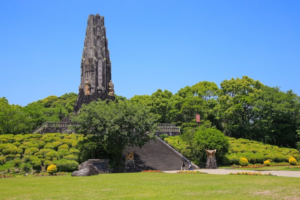 A large, ancient stone tower with a statue at its base, surrounded by lush green trees and a well-maintained garden.