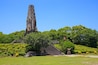 A large, ancient stone tower with a statue at its base, surrounded by lush green trees and a well-maintained garden.